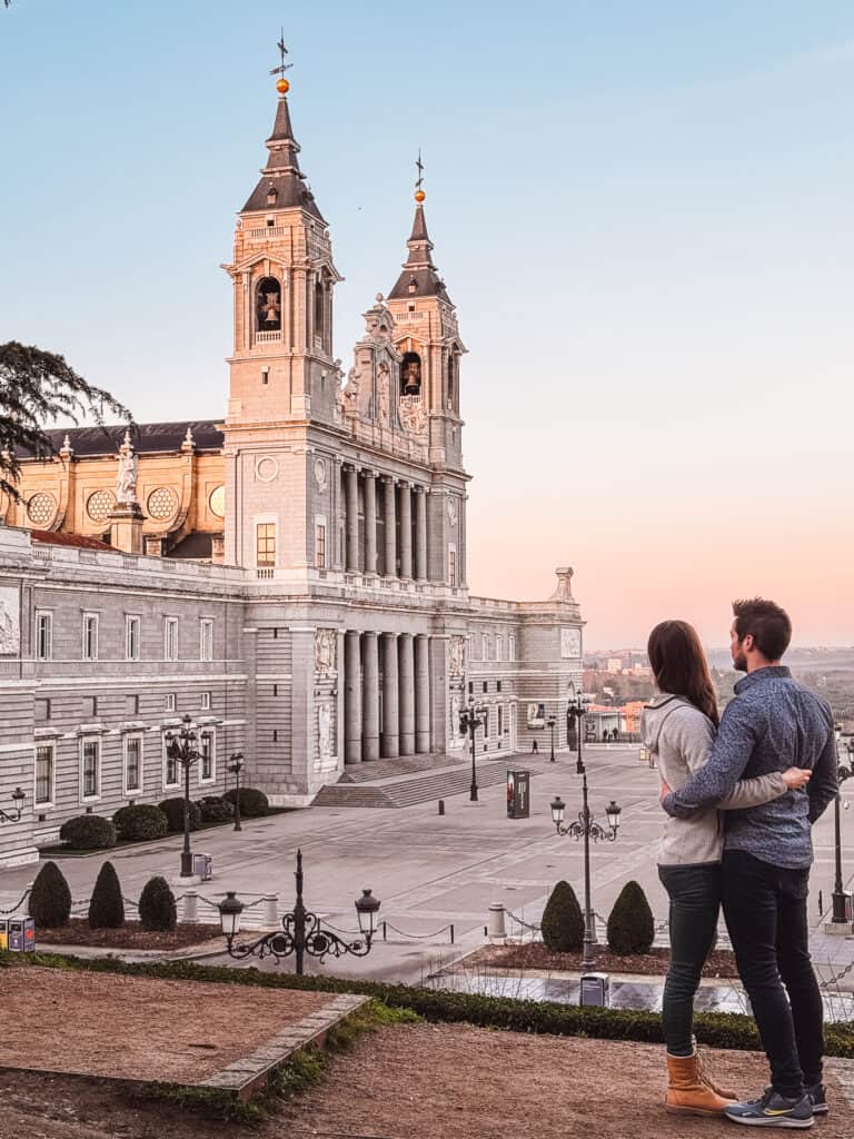 A couple stands embracing while looking at the Almudena Cathedral during sunset in Madrid, with the grand architecture and clear skies in the background