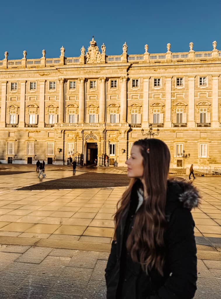 A woman in a black coat with long hair stands in the foreground, slightly out of focus, with the majestic Royal Palace of Madrid illuminated by the golden light of the setting sun in the background