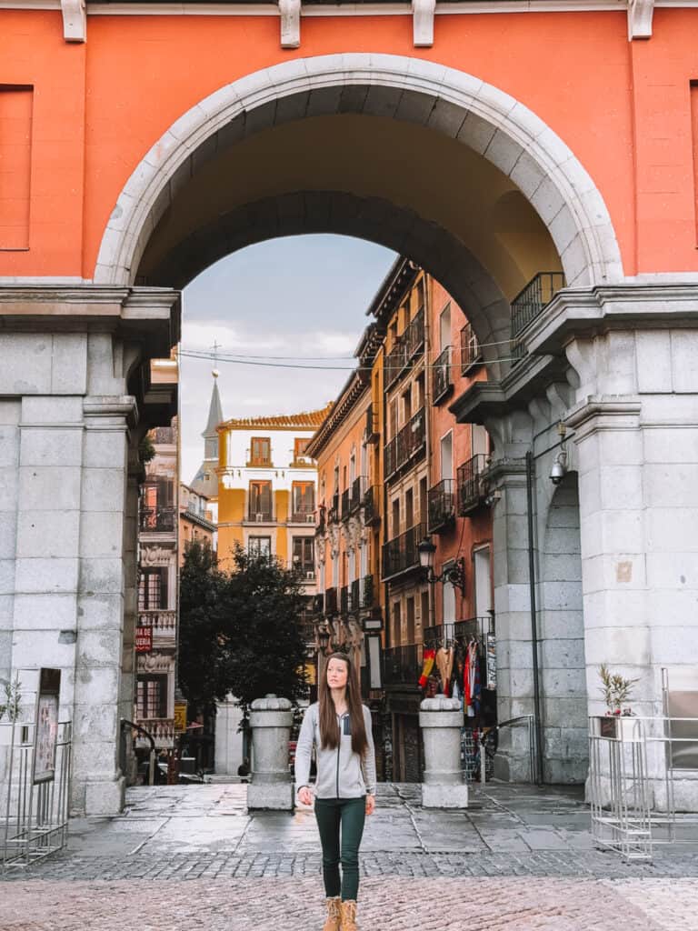 A woman in casual attire stands centered under a large archway, leading to a vibrant street with colorful buildings and balconies in Madrid, showcasing the city's lively architecture