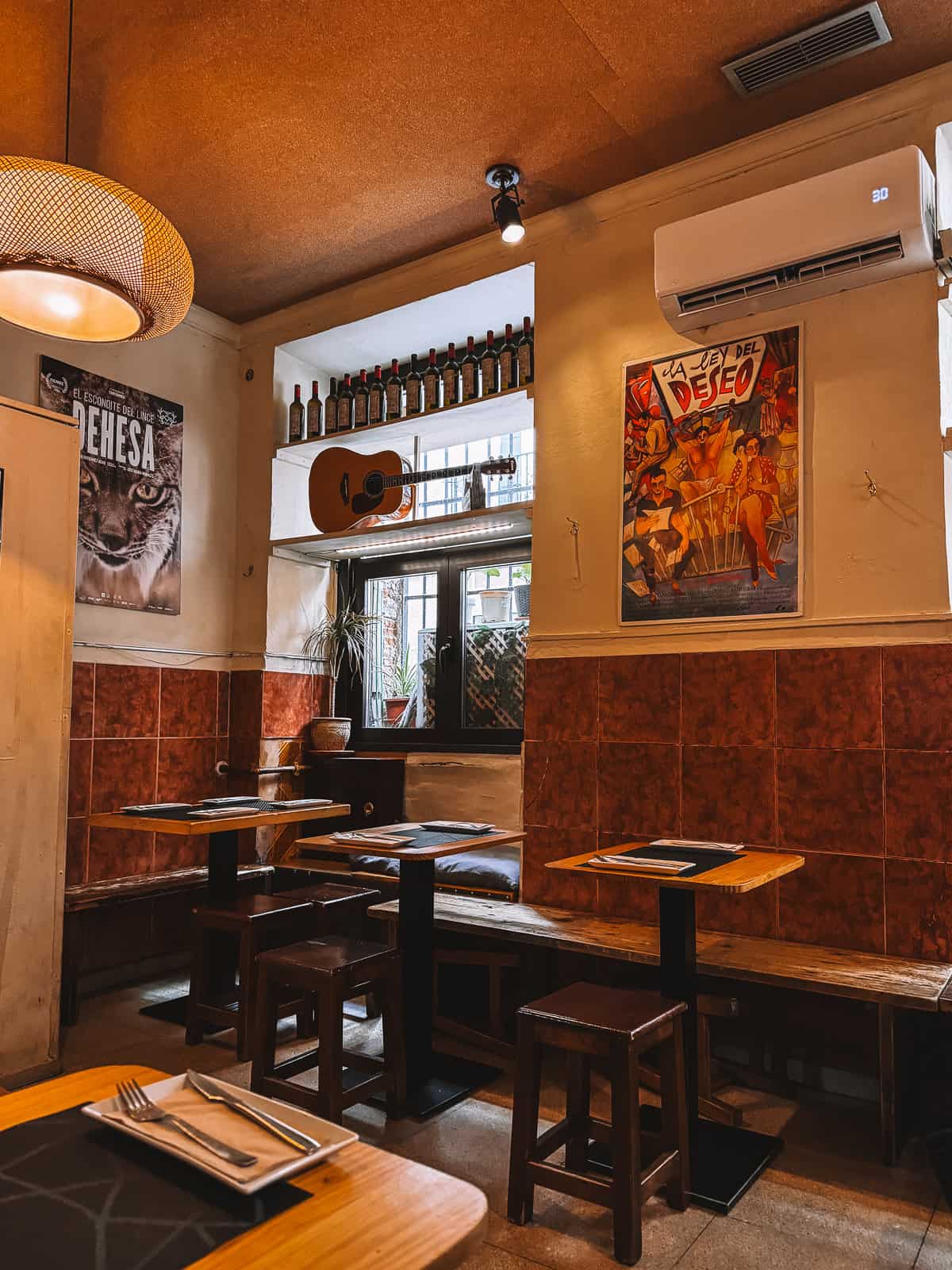 Cozy interior of 'Taberna El Sur' with wooden tables and benches, decorative bottles on a shelf, a guitar hanging on the wall, and a vintage Spanish movie poster