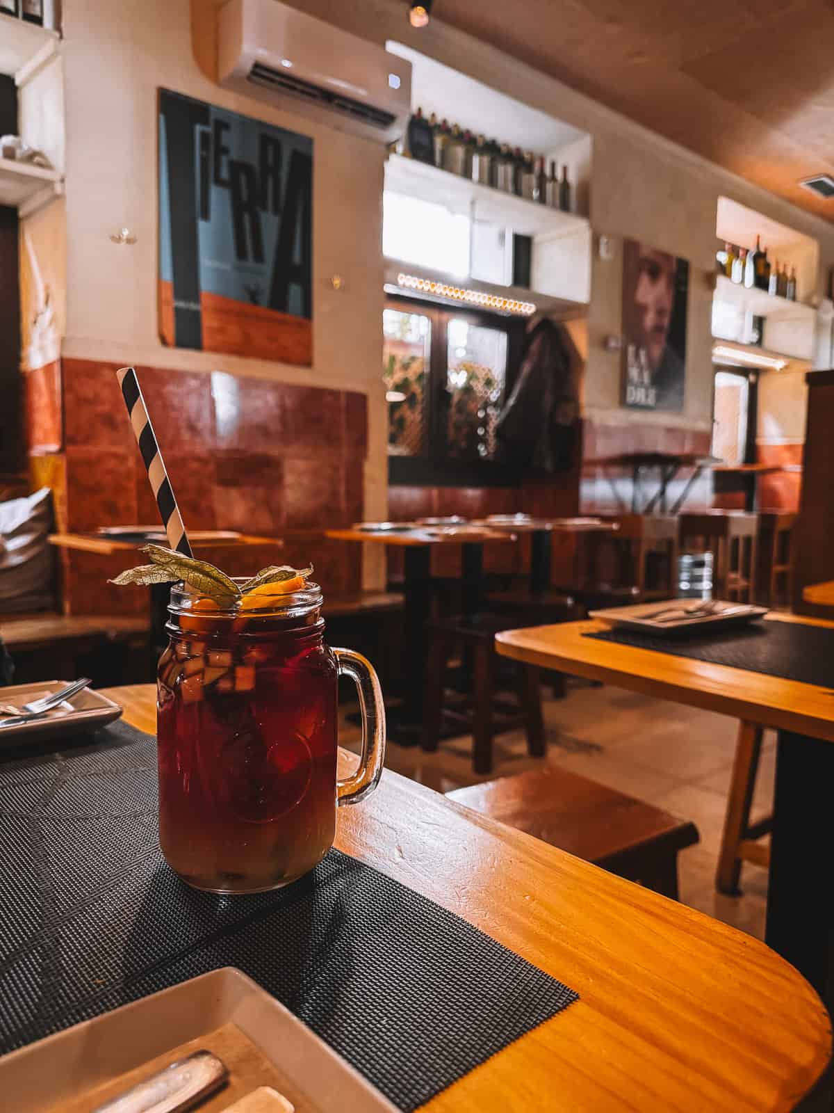 A refreshing sangria served in a mason jar with a striped straw, garnished with a slice of orange and lemon, placed on a wooden table inside the rustic 'Taberna El Sur'