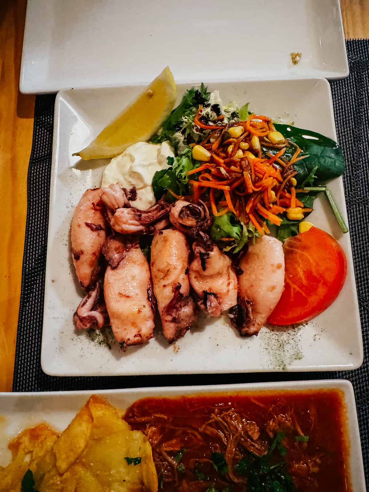 Plate of grilled calamari served with a lemon wedge, aioli, and a fresh salad, accompanied by a side of 'Patatas Bravas' in a cozy setting at 'Taberna El Sur'