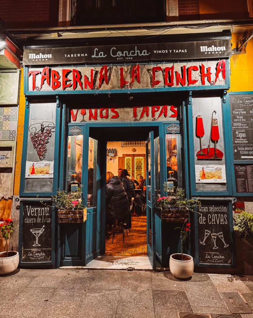 Exterior of 'Taberna La Concha,' a traditional Spanish tavern, invitingly open with patrons visible inside, framed by vibrant blue doors and decorative tiled facade.
