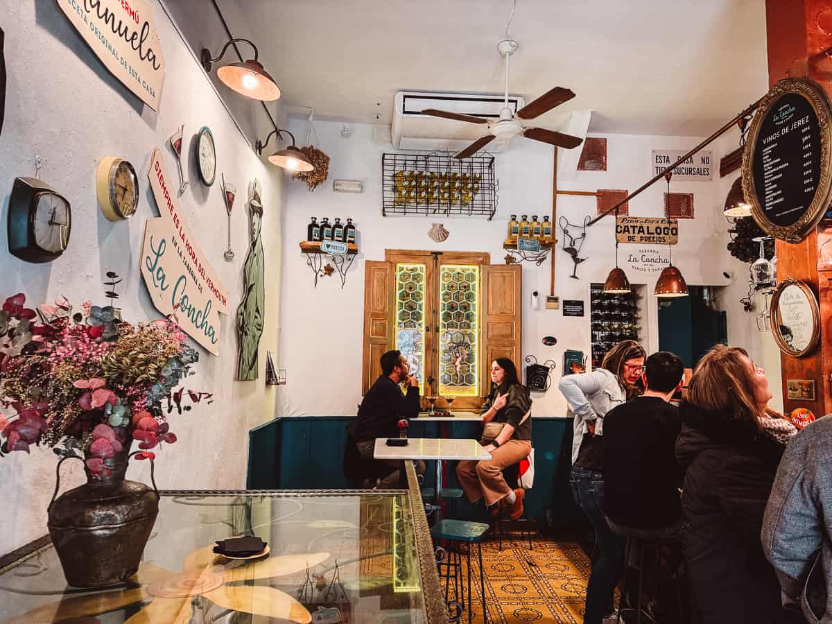 A cozy and vibrant tapas bar called Taberna La Concha interior with eclectic decorations, featuring wall-mounted plates, a variety of hanging glasses, and a large floral arrangement. Patrons are engaged in conversation, adding to the warm and social ambiance of the Spanish eatery.