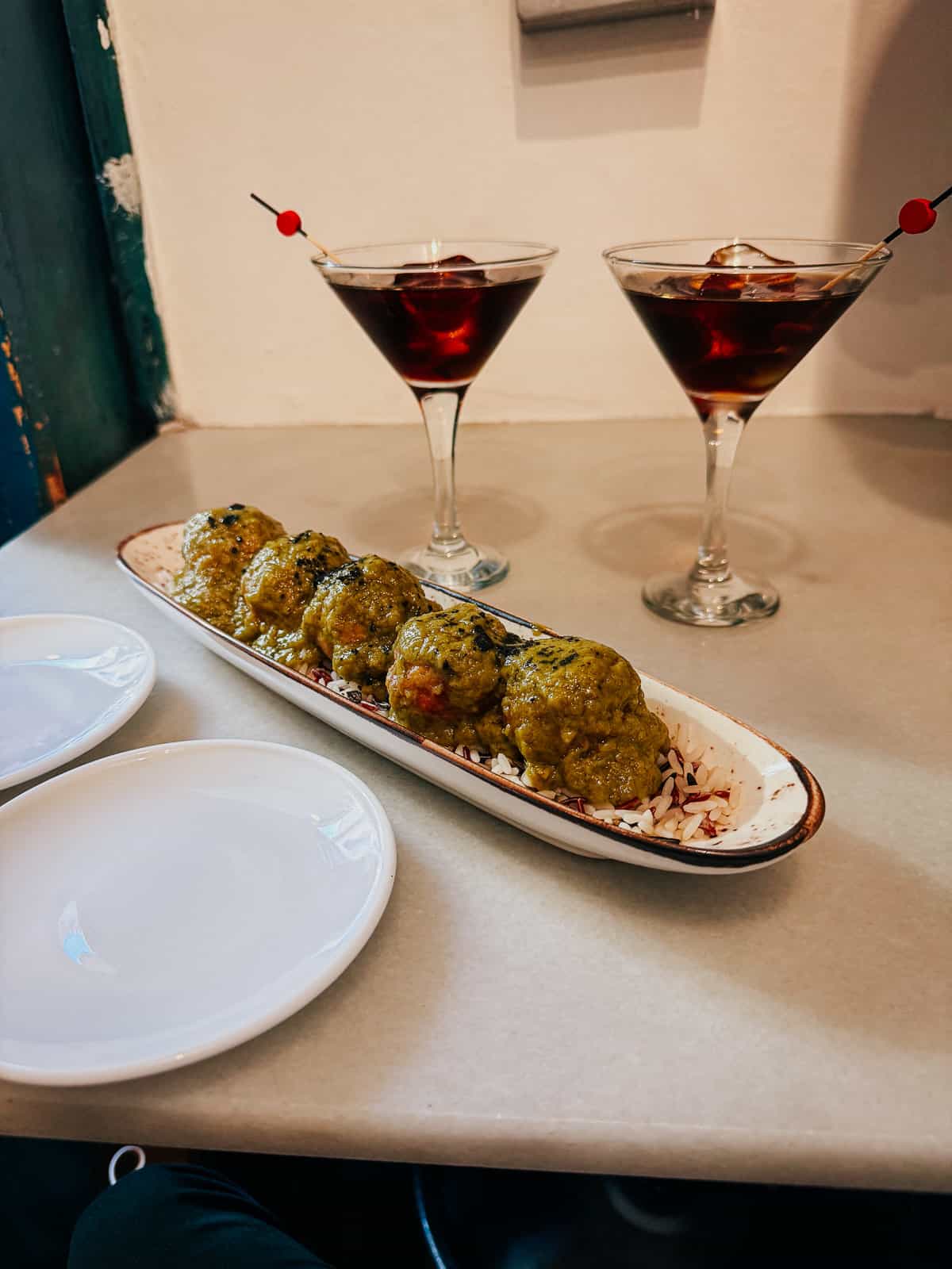 Dining table view at Taberna La Concha featuring a long oval plate of green, herb-coated meatballs next to two martini glasses with red vermouth, and empty white plates, suggesting a prepared dining experience