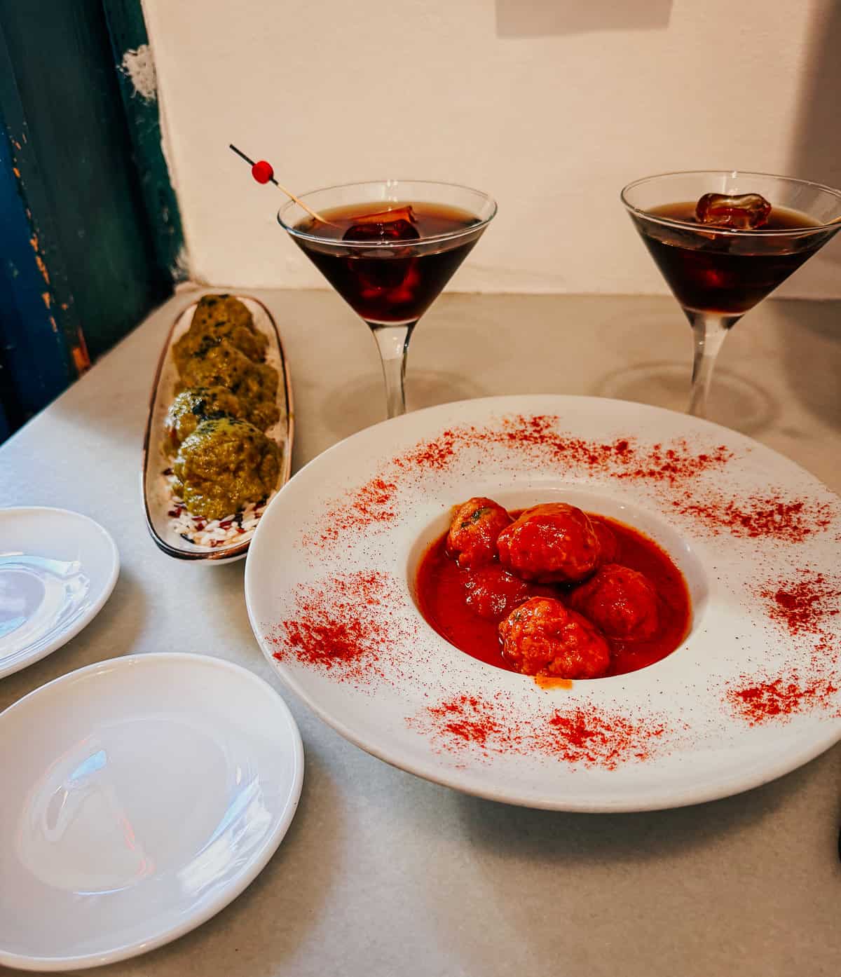 Dining table view featuring a long oval plate of green, herb-coated meatballs next to two martini glasses with red cocktails, and empty white plates, suggesting a prepared dining experience