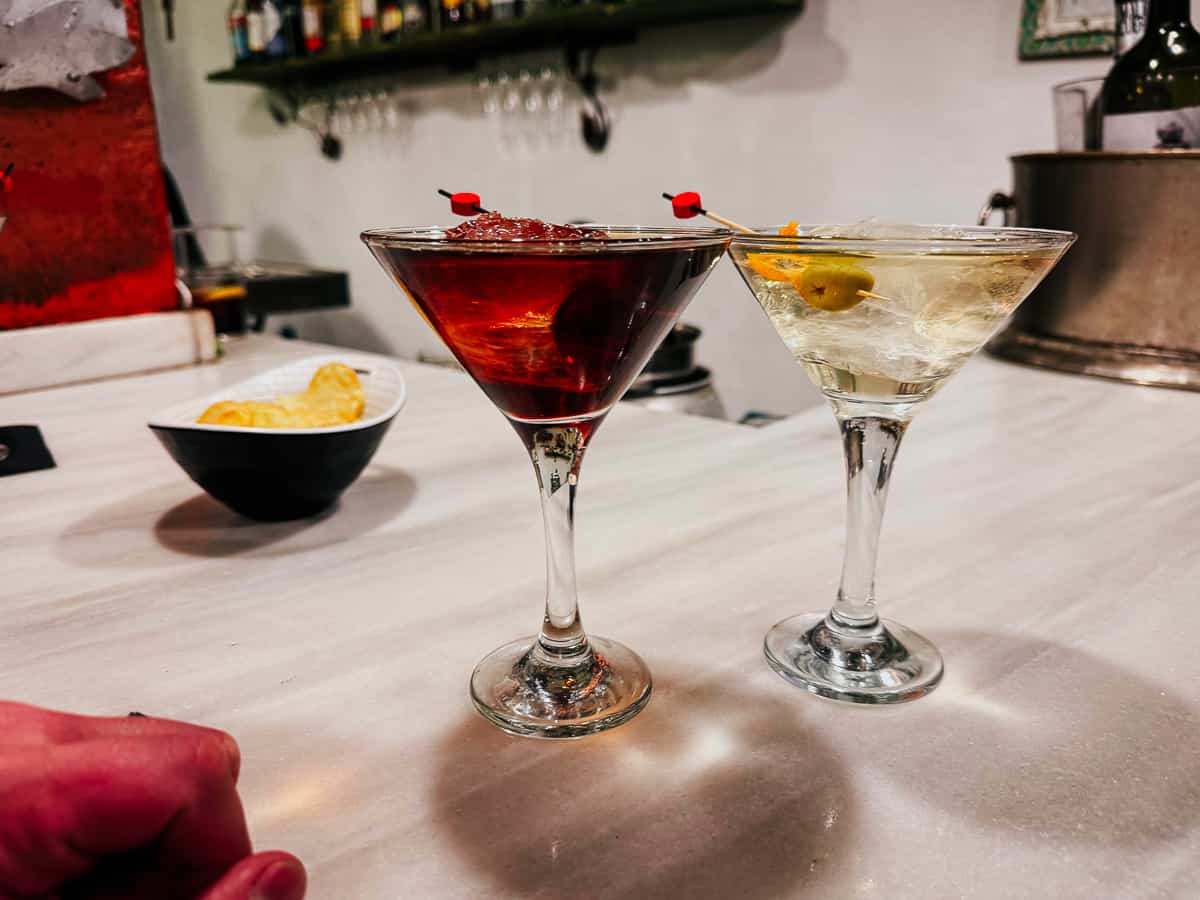 An intimate bar setting with two martini glasses, one with a dark vermouth garnished with a cherry, the other clear with an olive, on a white countertop, accompanied by a bowl of yellow crisps, suggesting a relaxed social atmosphere