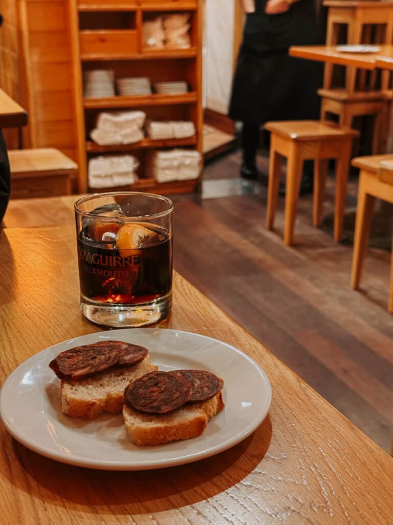 A rustic wooden table featuring a glass of vermouth accompanied by two toasted bread slices topped with a dark, cured meat, in a cozy bar setting with shelves in the background.