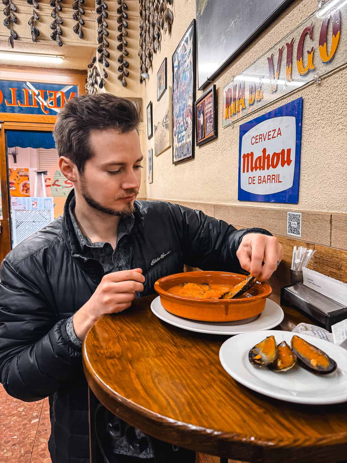A man in a black jacket is focused on eating a bowl of orange stew, with mussels on a side plate, at a bar decorated with hanging garlic strings and a Mahou beer sign.