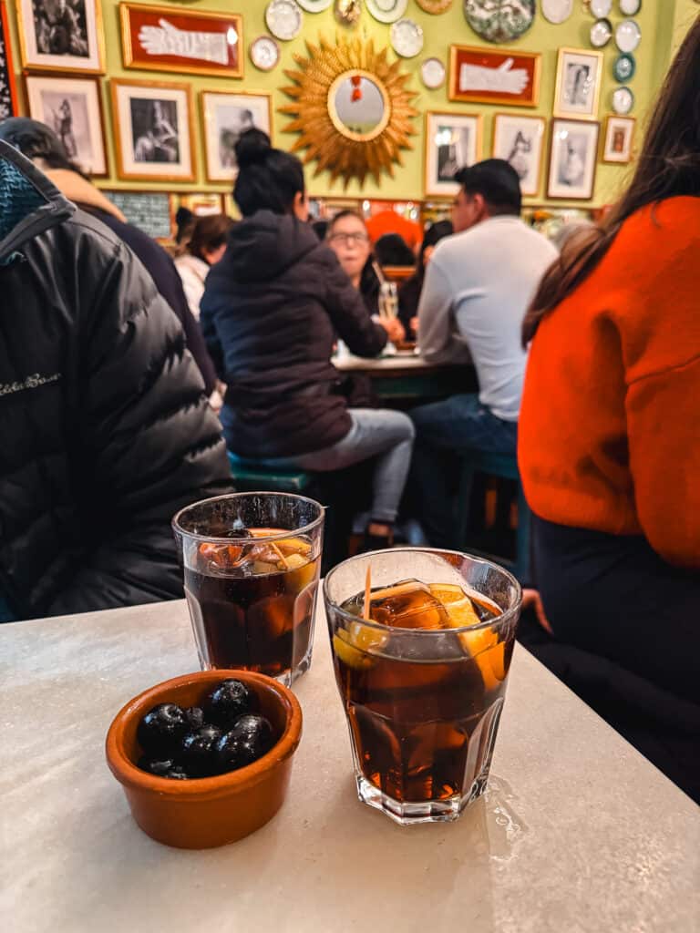Two glasses of vermouth on the rocks with a side of olives in a small earthenware bowl, on a marble tabletop in a lively bar with patrons in the background.