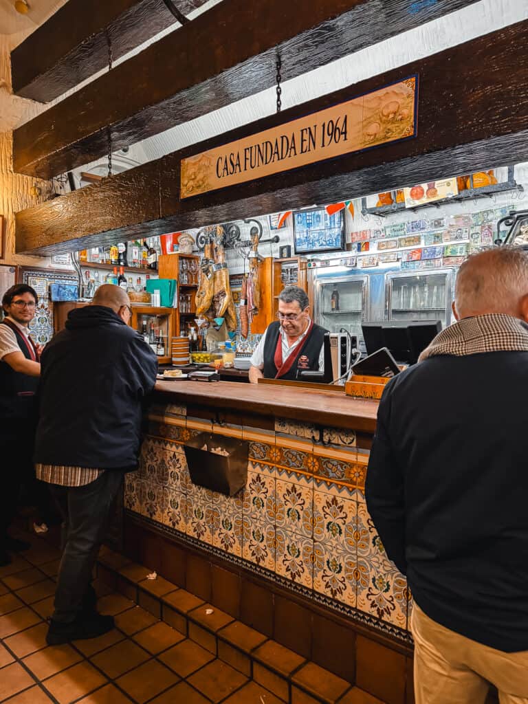 Interior of a traditional Spanish bar with ornate tiles, a wooden bar counter, patrons enjoying drinks, and a sign that reads 'Casa Fundada en 1964' indicating its establishment.
