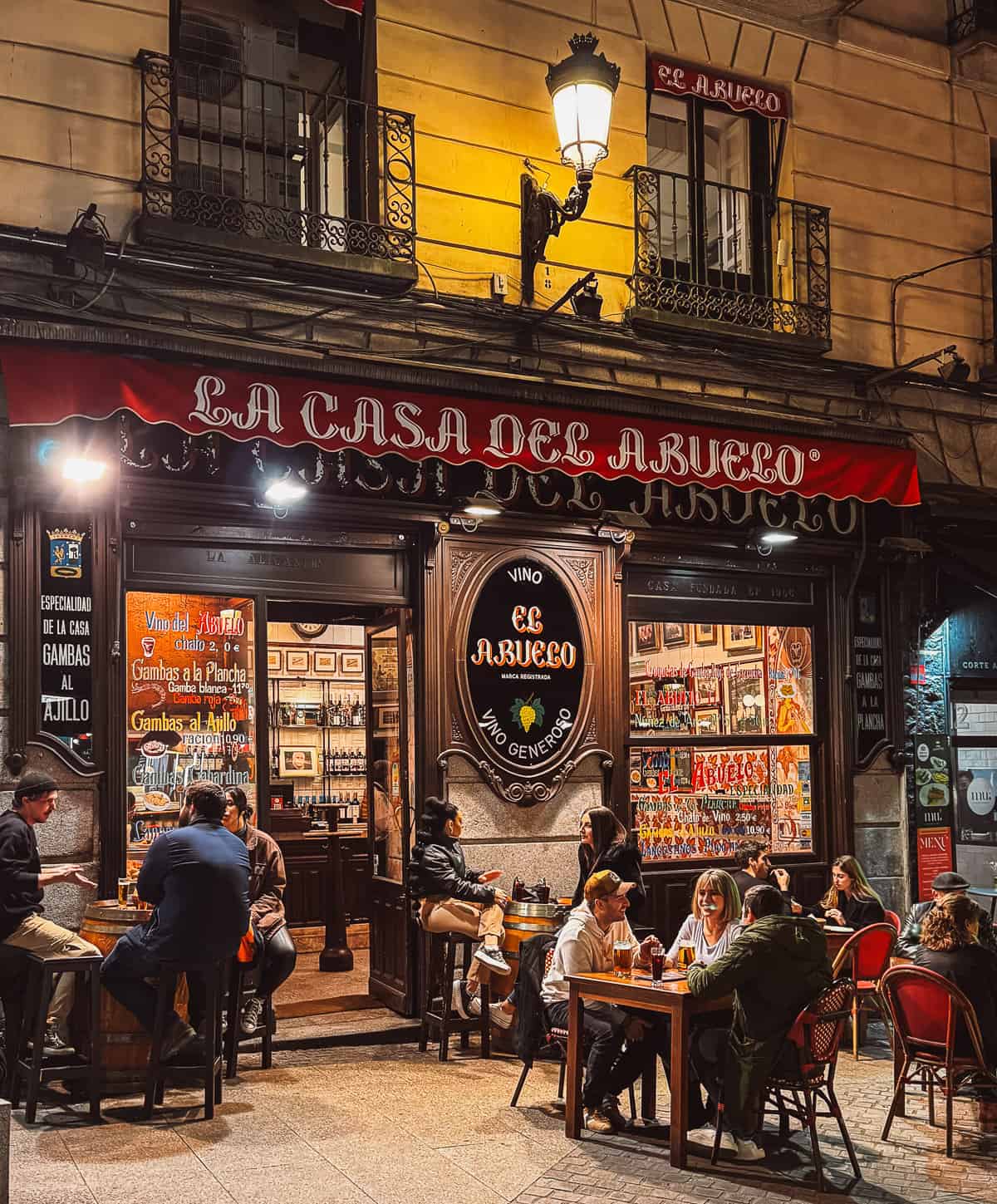 Evening view of 'La Casa del Abuelo', a traditional Spanish bar, with its warmly lit facade and patrons enjoying drinks at tables on the sidewalk under a quaint street lamp