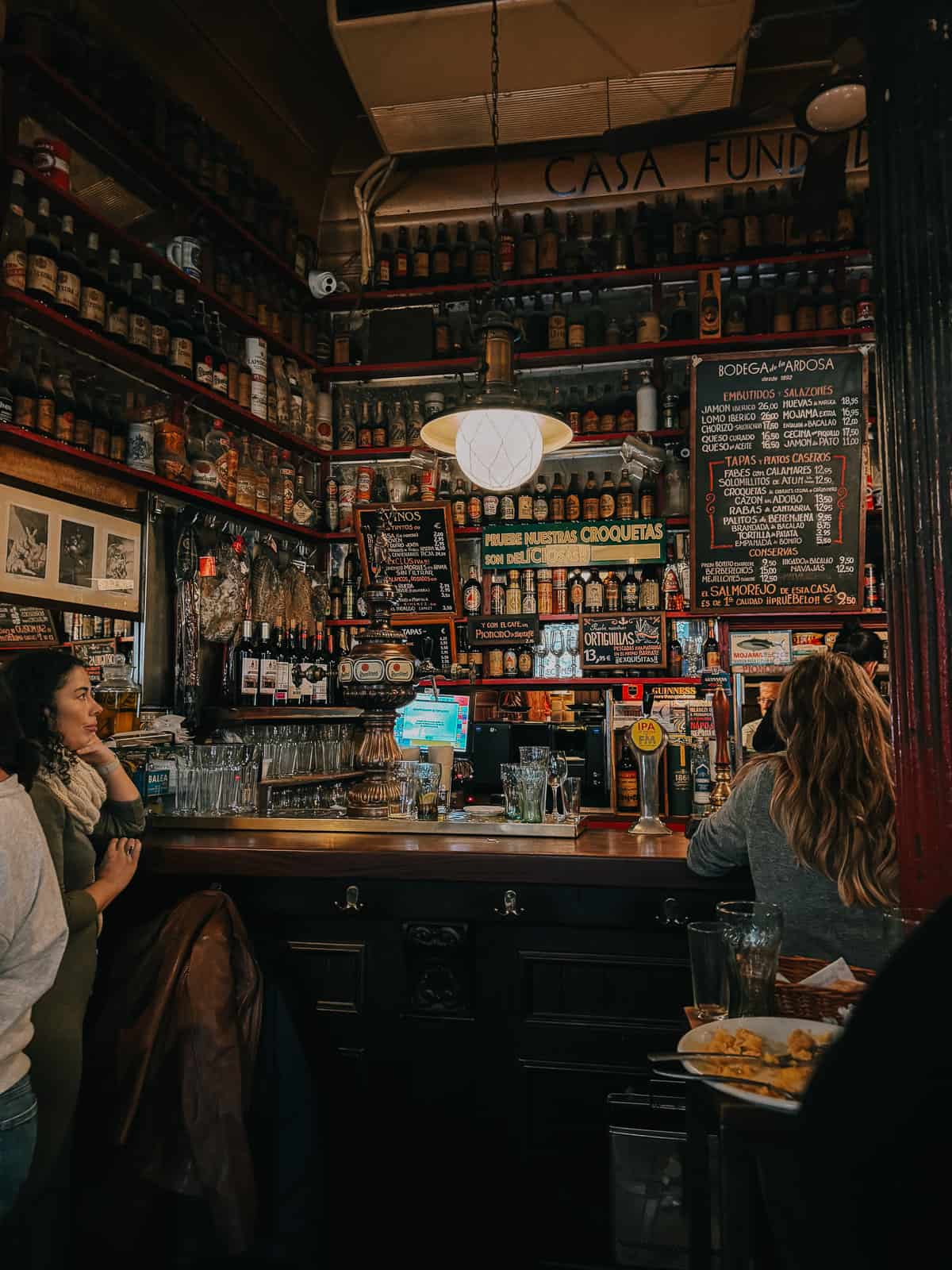 Interior of a classic Spanish bar with a variety of wines and spirits lining the shelves, a detailed menu board, and customers engaged in conversation by the wooden bar counter