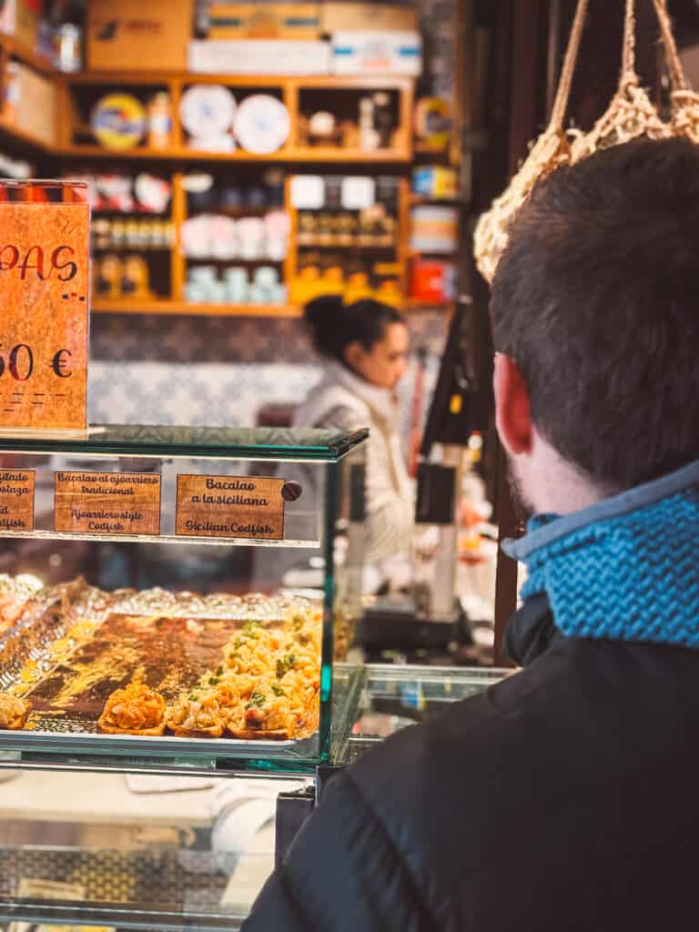 A customer from a rear view looking at a glass counter filled with trays of 'tapas', showcasing a variety of Spanish appetizers, inside a bustling tapas bar.