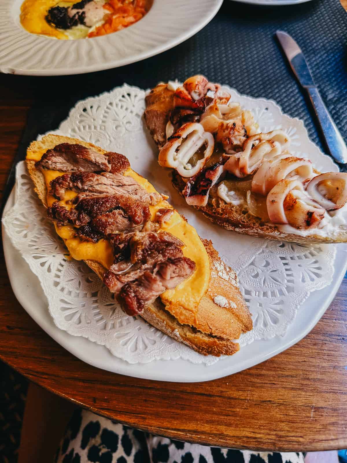 Open-faced sandwiches on crusty bread, one topped with melted cheese and chicken, the other with cheese and calamari rings, served on a lace paper doily