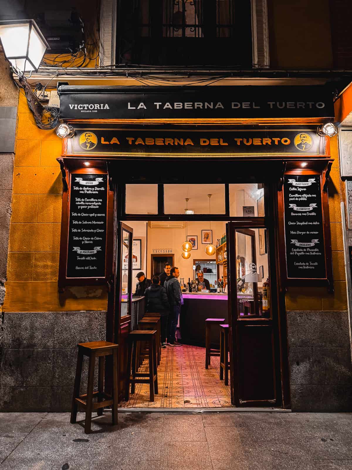 The entrance of 'La Taberna del Tuerto' at night, showcasing its warm yellow and purple facade, inviting patrons inside under a lit signboard