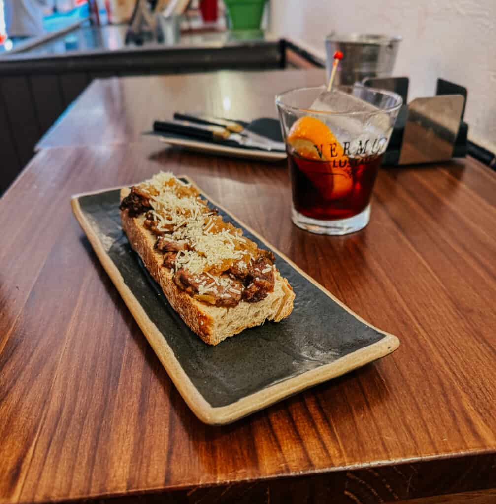 A rustic tapa of crusty bread topped with pork cheek and grated cheese, accompanied by a glass of vermouth, on a wooden bar counter.