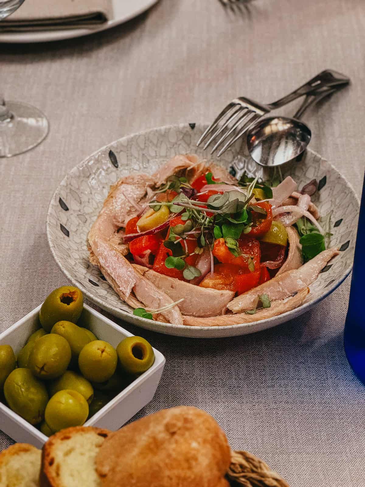 A fresh tuna salad with vibrant tomatoes, green olives, and microgreens served in a glass bowl on a textured tablecloth, accompanied by bread and a bowl of olives, ready for dining