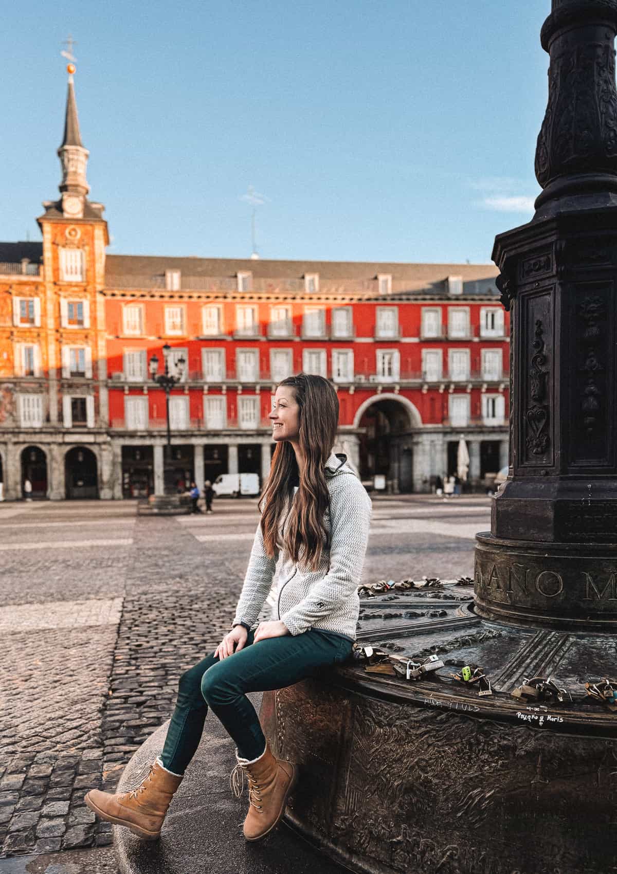 A woman sits contentedly at the base of an ornate lamppost in Plaza Mayor, Madrid, with the square's iconic red facades and arched portals in the background, while love locks adorn the post, symbolizing the romance of the city.