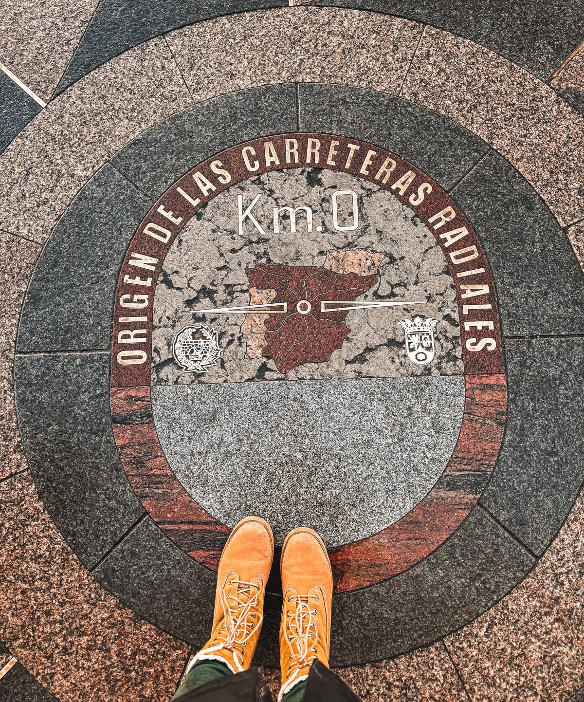 A pair of feet in bright orange shoes stand atop the Km 0 marker in Puerta del Sol, Madrid, the central point from which all distances in Spain are measured, indicating the historic and geographic heart of the country.