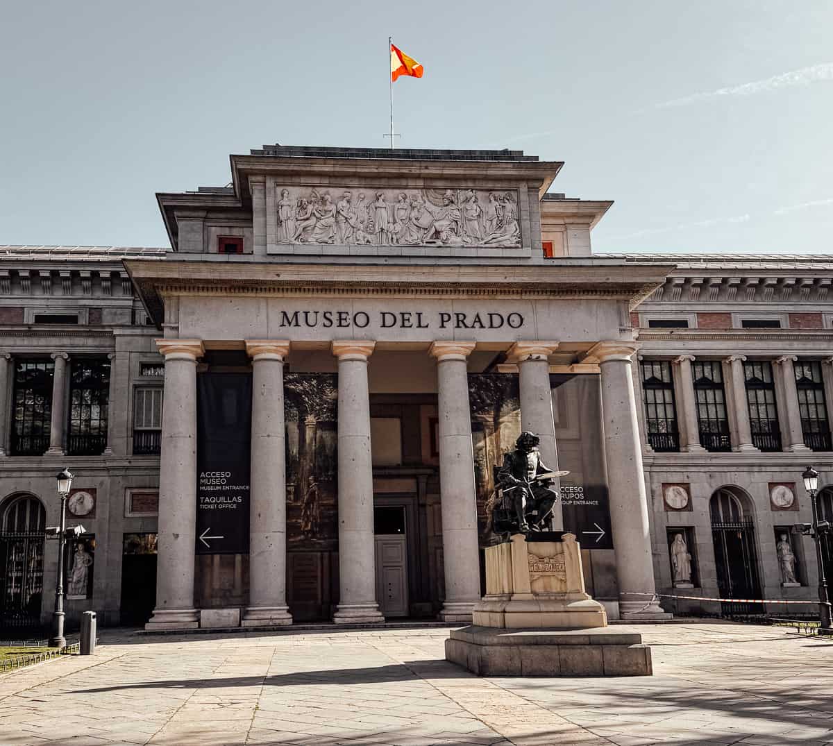 The neoclassical facade of the Museo del Prado in Madrid, with its towering columns and the Spanish flag waving above, representing one of the most significant cultural landmarks in Spain.