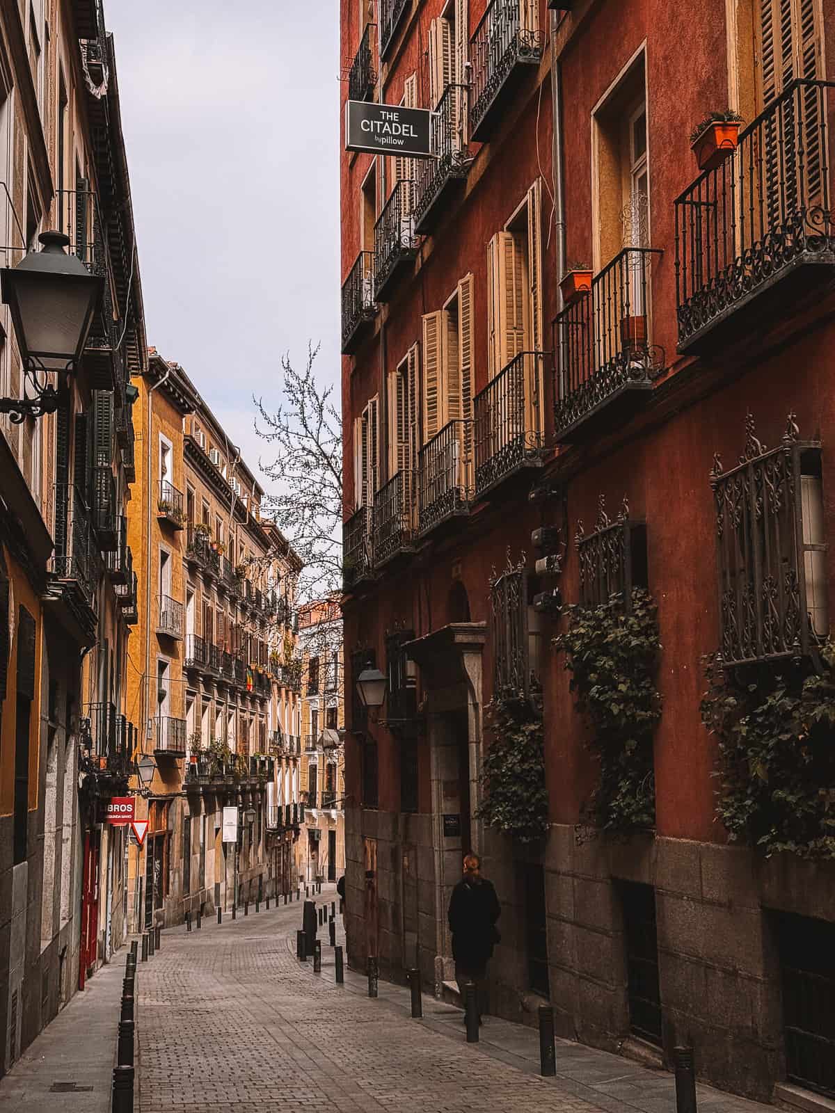 A quiet, cobblestone street in Madrid flanked by traditional buildings with wrought-iron balconies and vibrant facades, exuding the historic charm of the city's old quarters.