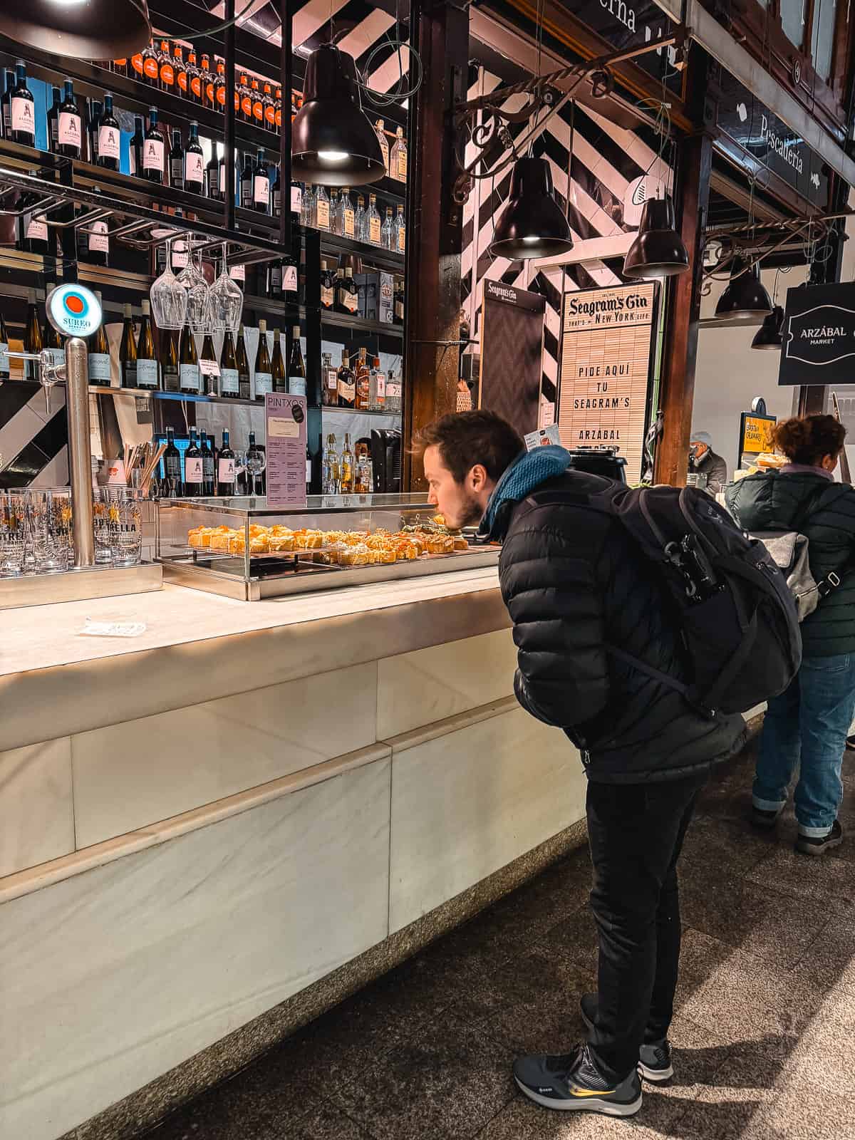 A patron peruses the offerings at a bustling market bar, with a variety of gin bottles on display above and an array of tapas in the foreground, showcasing a slice of local gastronomy and nightlife