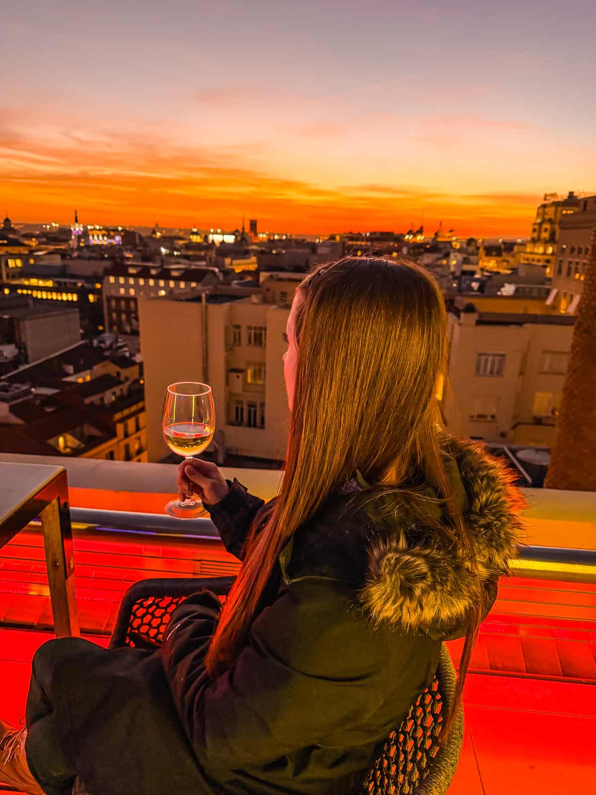 A woman enjoys a glass of wine on a rooftop terrace at sunset, with a panoramic view of Madrid's cityscape bathed in warm hues of orange and red, reflecting a tranquil urban evening