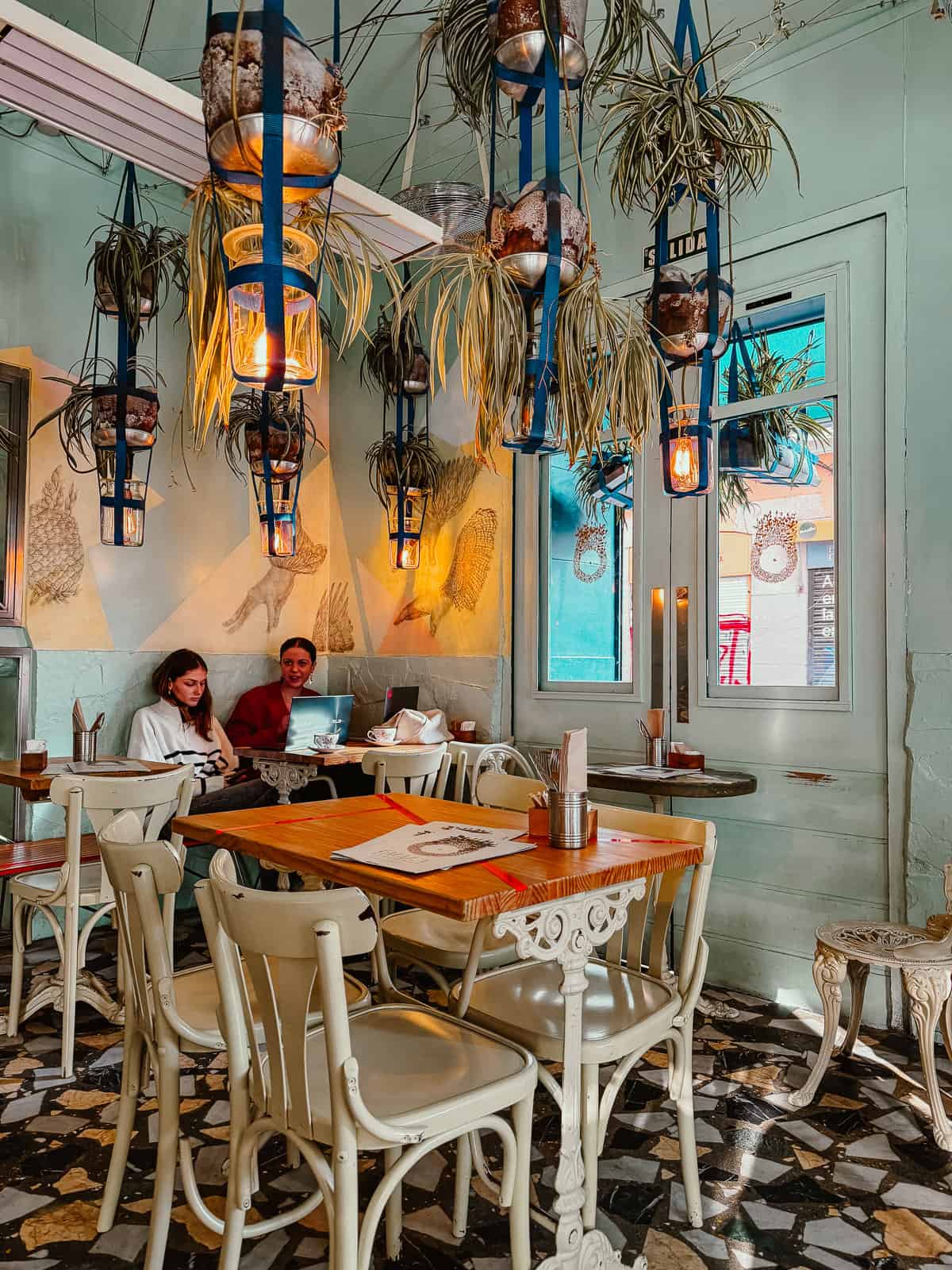 A cozy, eclectic café interior with hanging potted plants and unique lanterns, where patrons engage in conversation by a window with a view to the street, surrounded by vintage-style furniture and artistic wall illustrations