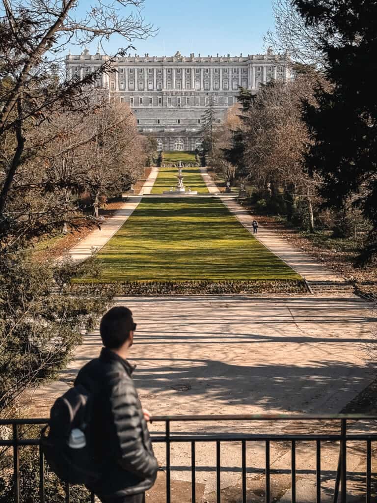 A person gazes out over the manicured hedges and pathways of the Sabatini Gardens, with the grand Royal Palace of Madrid in the distance, all under a clear blue sky