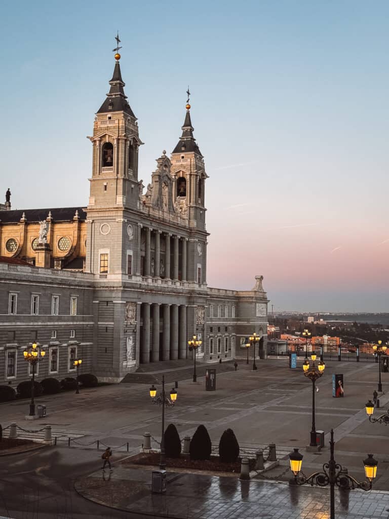 The Almudena Cathedral stands majestic during twilight in Madrid, its baroque architecture illuminated against the fading sky, while the Plaza de la Armería below glows with street lamps and stretches out serenely