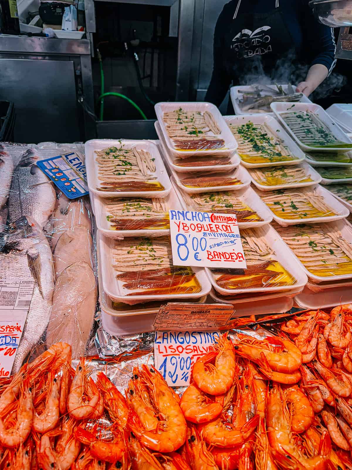 A seafood stall displays fresh anchovies in vinegar and succulent langoustines with prices labeled, providing a colorful and appetizing glimpse into the culinary delights offered at a Spanish market