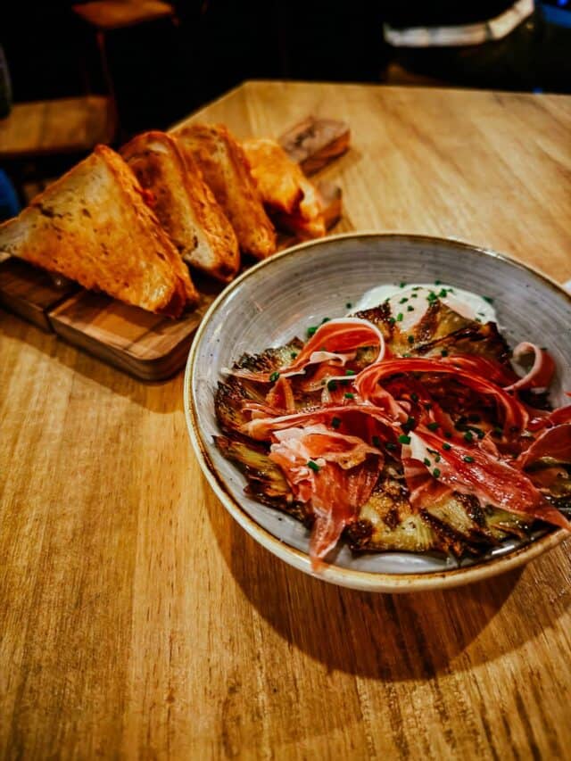 A rustic wooden table with a dish of grilled artichokes topped with thin slices of jamón ibérico, accompanied by a serving of toasted bread, exemplifying a traditional Spanish tapas meal.