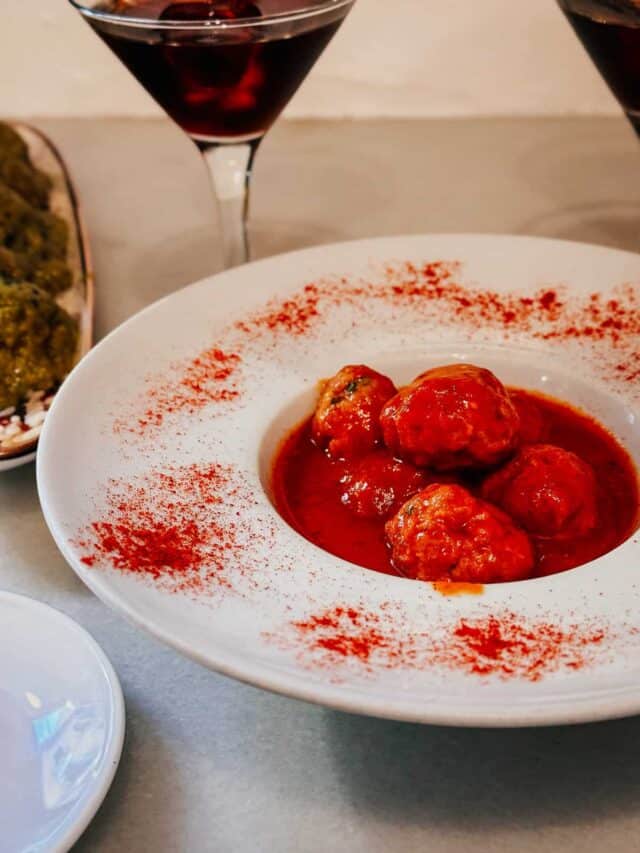 Close-up of a plate of meatballs in a rich red sauce, dusted with a sprinkle of spices, paired with two dark red martinis, showcasing an appetizing meal setup.
