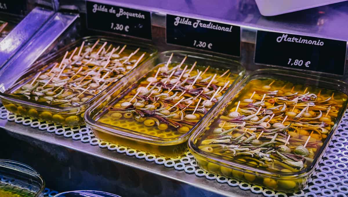 Gilda de Boquerón and traditional skewered Gilda and Matrimonio appetizers submerged in olive oil, priced at 1.30€ and 1.80€ respectively, displayed in a Spanish tapas bar