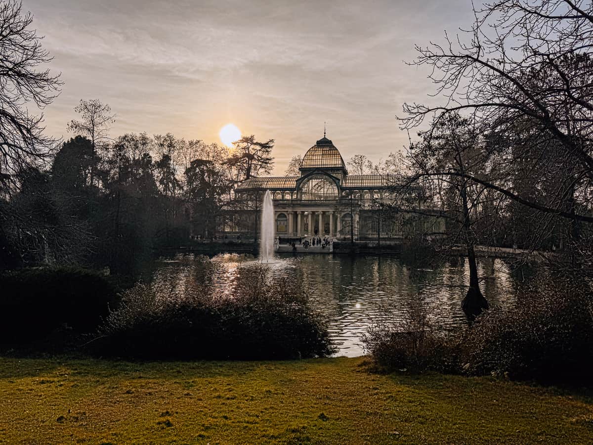 Sunset view of the Crystal Palace in Madrid's Retiro Park, with the sun casting a soft glow over the structure and reflecting on the calm pond surrounded by lush greenery.