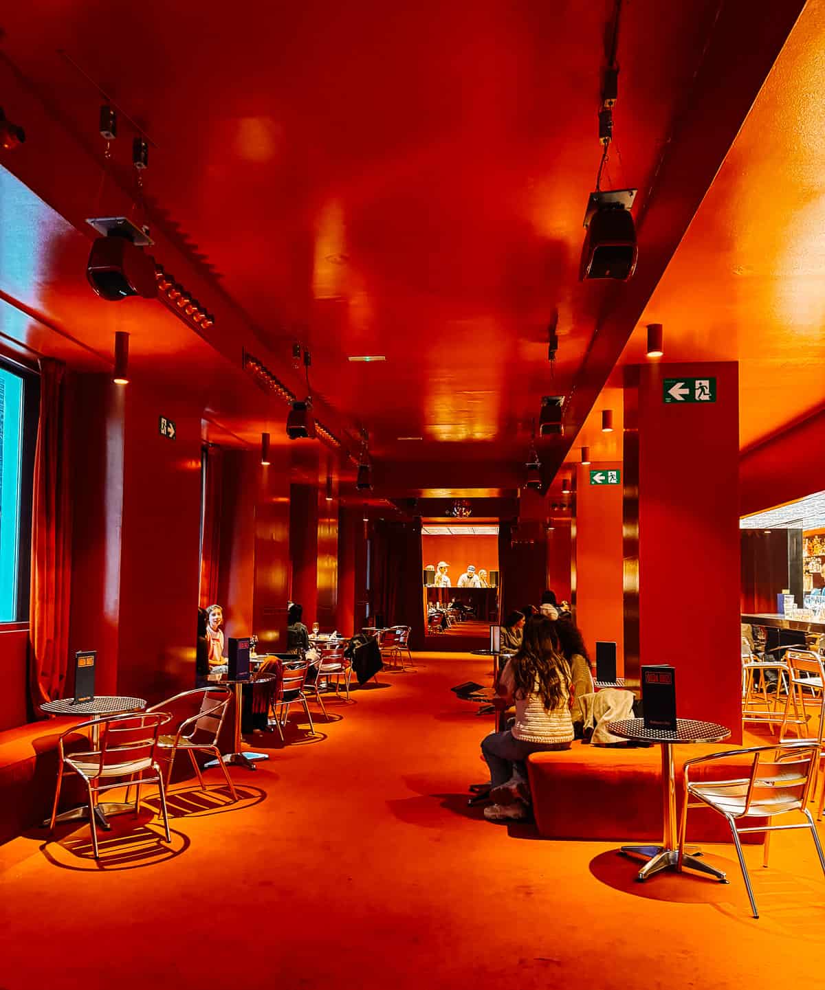 Vibrant red lounge area with contemporary decor, featuring a line of tables and chairs along a corridor leading to a stage with a digital display, creating an inviting modern space for socializing.