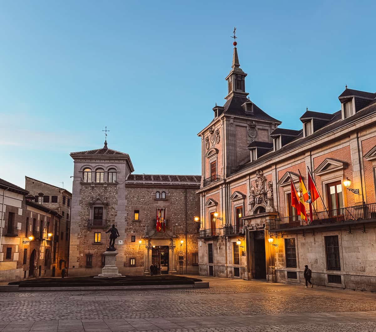 Twilight over Plaza de la Villa, one of the hidden gems in Madrid, featuring historic buildings adorned with Spanish flags under a clear sky.