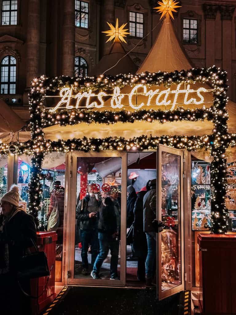 An illuminated 'Arts & Crafts' sign adorned with twinkling lights at a festive outdoor market booth, with shoppers visible through the glass door and a star decoration hanging above.