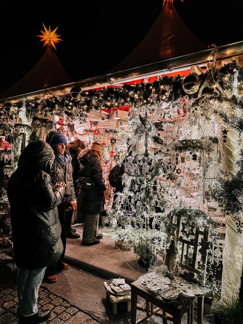 Visitors browsing an array of intricately designed snowflake decorations at a festive market stall, with sparkling lights and Christmas greenery enhancing the cozy holiday atmosphere.