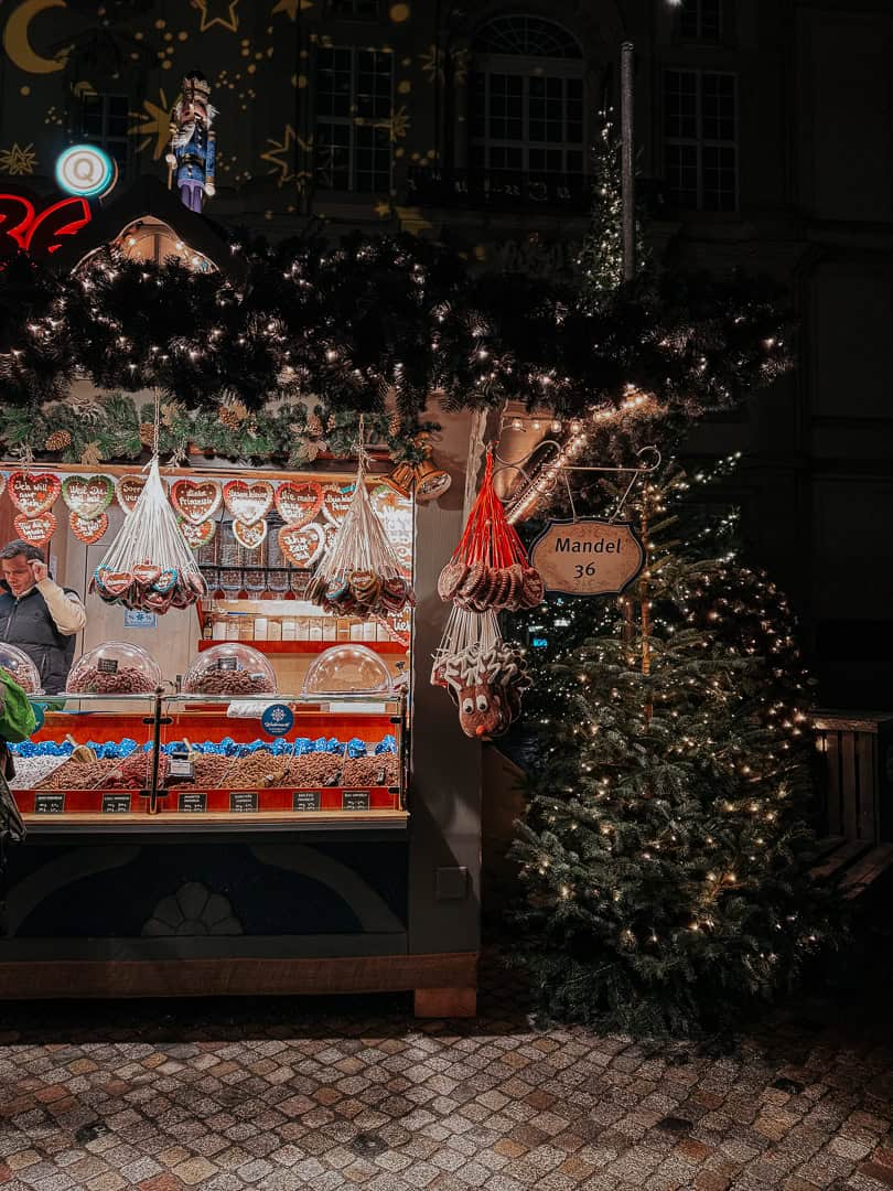 A traditional Christmas market stall adorned with lights and a variety of gingerbread hearts hanging on display, next to a sign for 'Mandel' and a festively decorated fir tree.