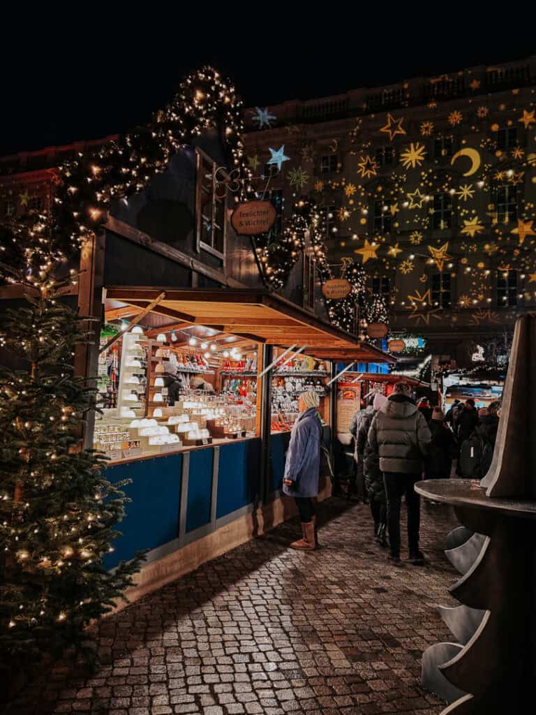 A vibrant Christmas market scene with a cobblestone pathway leading to a warmly lit market stall under a festively decorated tree, with a large building illuminated with projected stars in the background.