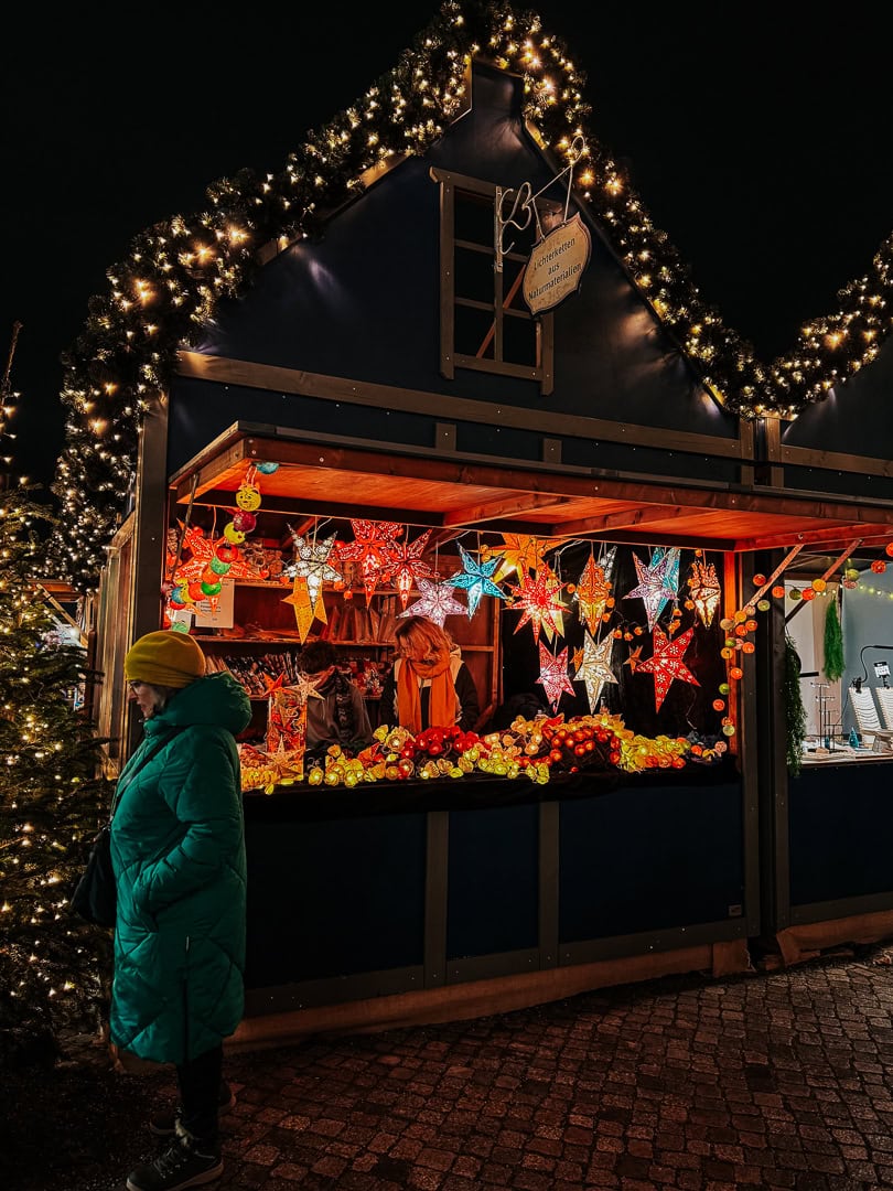 A cozy Christmas market booth under a twinkling garland roof, selling colorful star-shaped lanterns and decorations to cheerful visitors in winter attire.