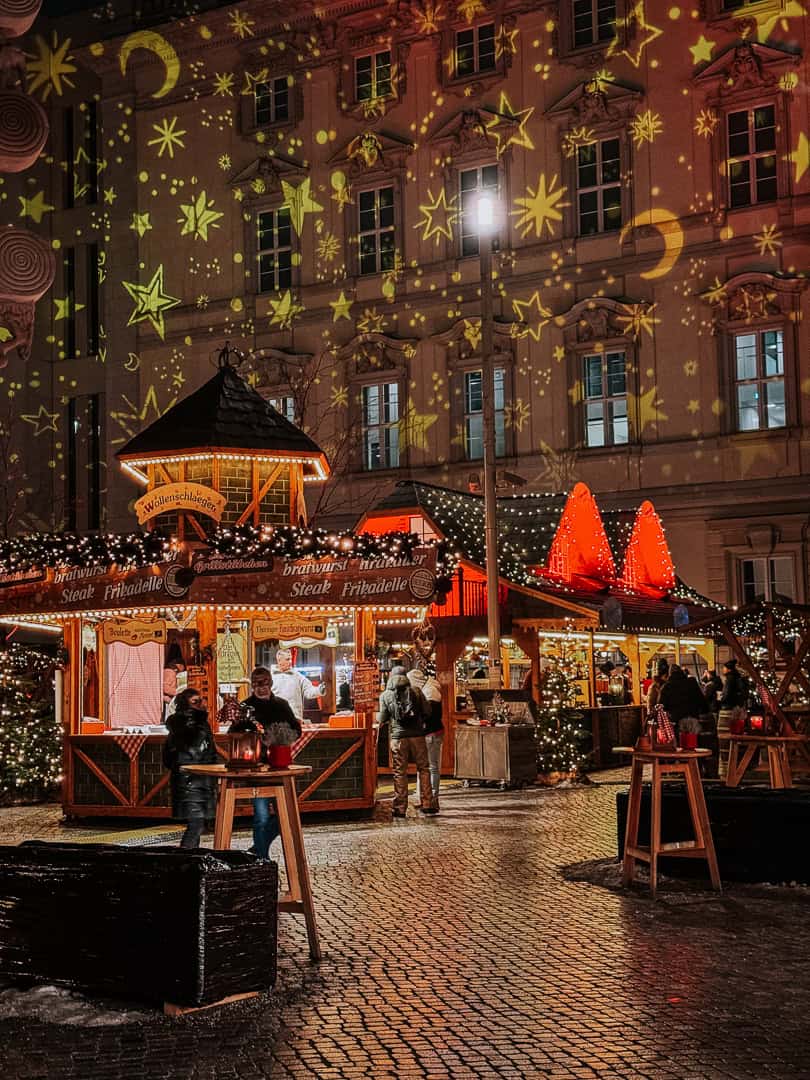 Evening at a Christmas market showcasing a food stall with 'Bratwurst', 'Steak', and 'Frikadelle' on offer, nestled under a building with a starry light projection.