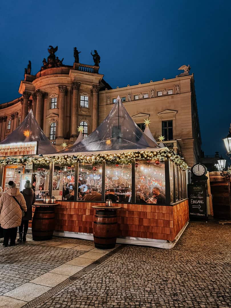 Patrons inside a warmly lit, tented pavilion at a Berlin Christmas market with festive decorations and a historic building in the background