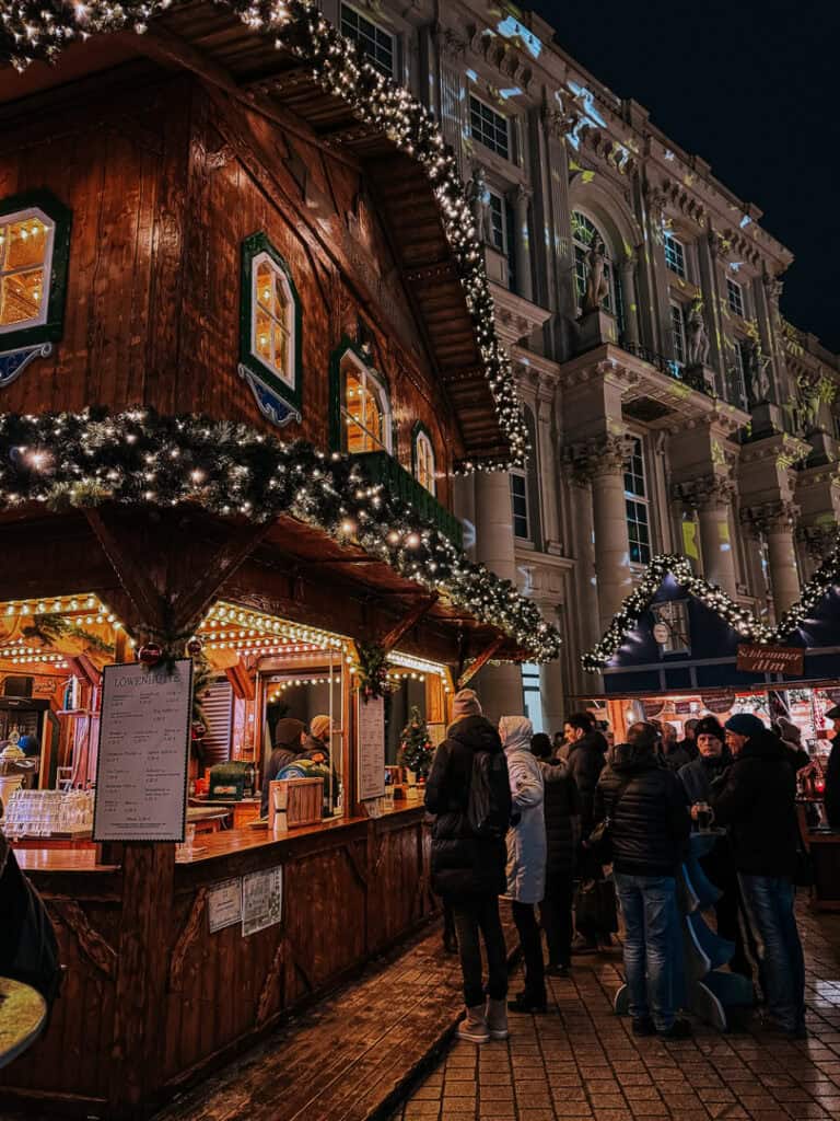 A festive wooden Christmas market booth festooned with twinkling garland lights, with people queueing up to place their orders, against the backdrop of a grand illuminated building.