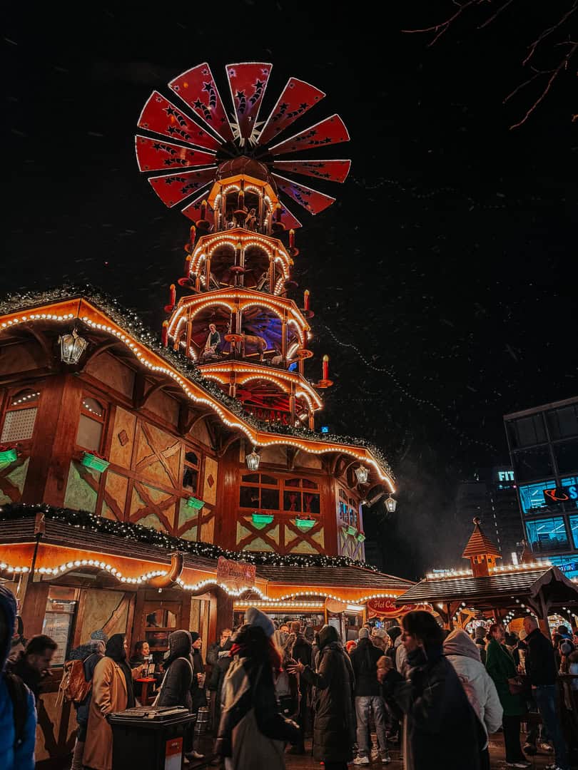 A majestic multi-tiered Christmas carousel with a large red star on top, surrounded by bustling market-goers and lit up against the night sky.