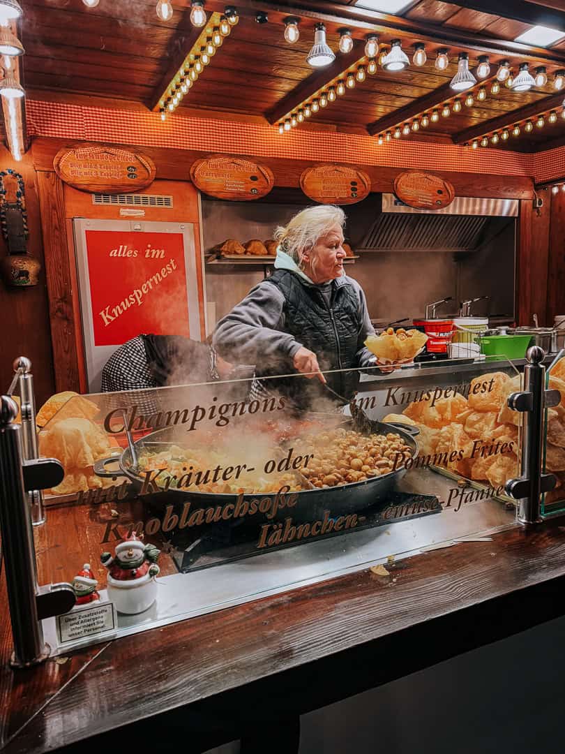 A vendor at a Christmas market serves steaming hot Champignons from a large pan, beneath a sign detailing the menu options, with festive decorations surrounding the cozy stall.
