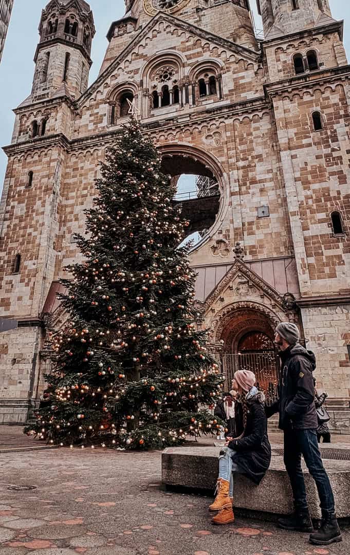 A large Christmas tree beautifully decorated with lights and ornaments stands in front of a historic church, with people sitting and standing around it, enjoying the holiday scene.