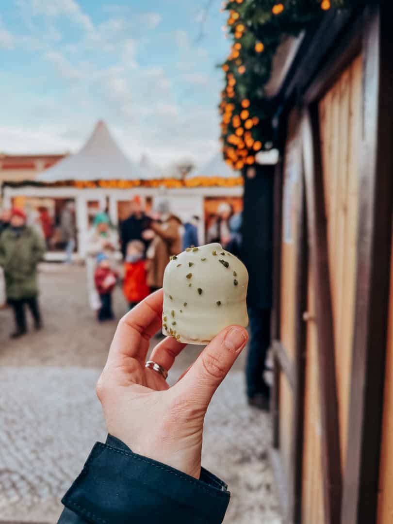 A close-up of a hand holding a white chocolate-covered marshmallow sprinkled with green pistachios, with a blurred background of a Christmas market.